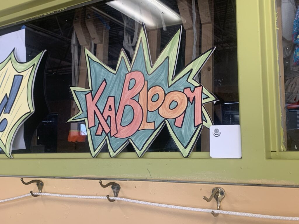 A white Conserv sensor placed in the corner of a window with lime green trim with a cutout painted artwork with the word, "Kabloom"