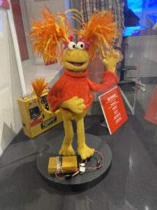 Yellow Fraggle puppet wearing an orange sweater. It has orange and yellow pompon tails on her head and tail.
