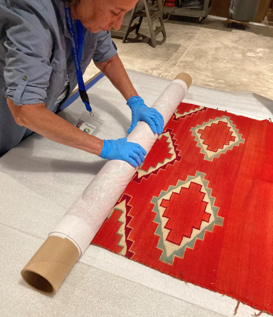 Rolled textile storage at the Arizona Museum of Natural History Anthropology volunteer Susan rolling a Navajo serape blanket. Woman with blue nitrile gloves leans over a red Navajo serape blanket as she rolls it into a tube. Image courtesy of the Arizona Museum of Natural History.
