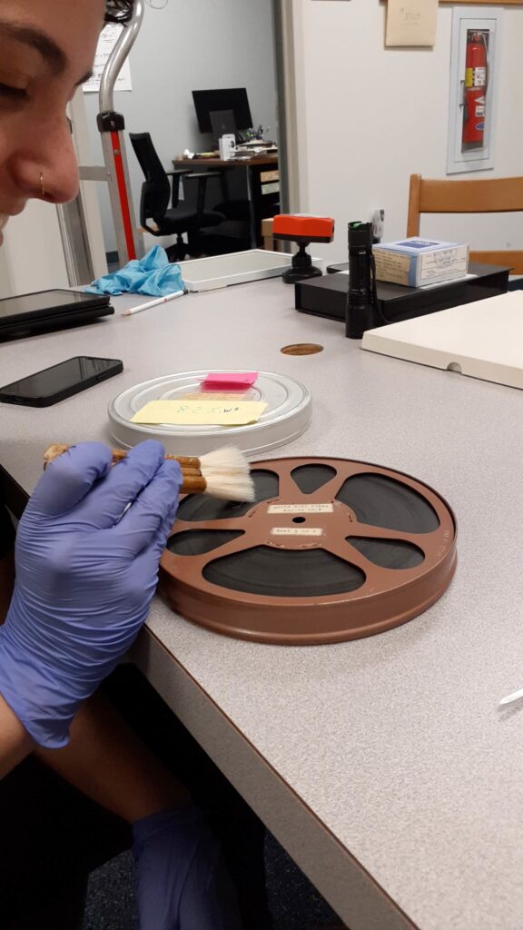 Collections Assessment for Preservation at the Disaster Research Center - A person with purple nitrile gloves using a soft specialised brush to surface clean a film reel.