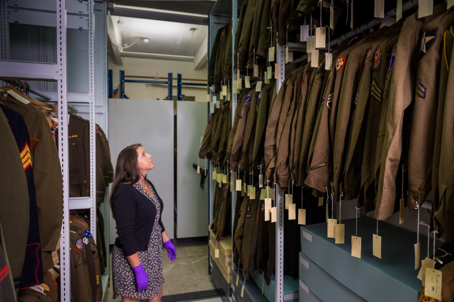 Remote Environmental Monitoring Equipment for Museums With The National WWII Museum Toni Kiser standing in storage spaces. In front, collection of WWII jackets with tags.