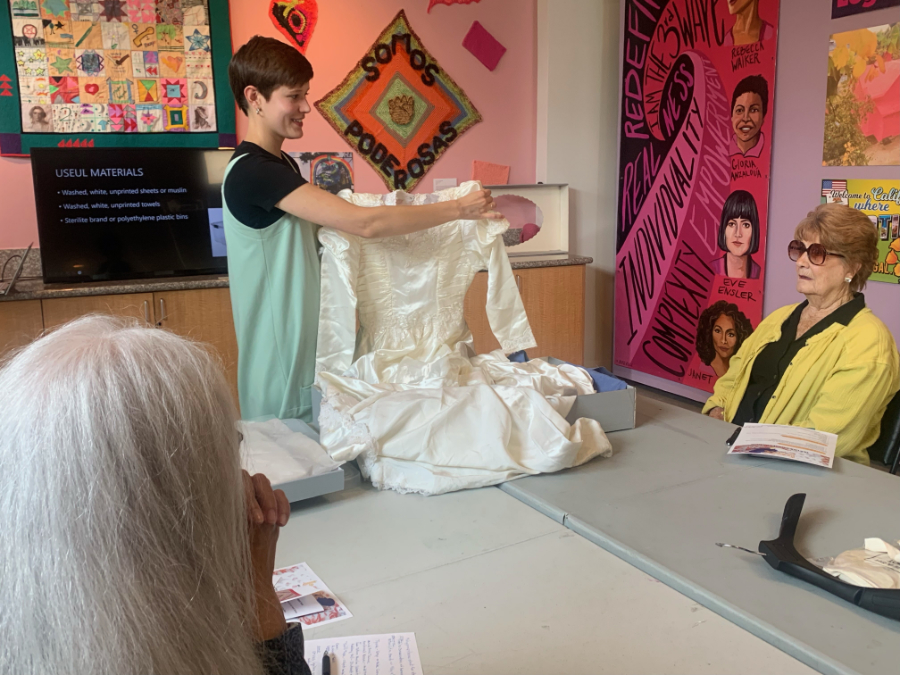 Woman in black T-shirt and teal dress holding up a wedding dress over a table. Two women sitting at the table watch the presentation.